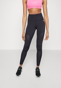 Zwarte high-waisted legging met een gladde textuur, voorzien van geometrische paneelontwerpen en gecombineerd met zwarte sportschoenen.