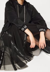 Czarna bluza z kapturem z błyszczącym sznurkiem, zestawiona z czarną spódnicą z tiulu z ozdobnymi taśmami z logo. Całość dopełniają czarne trampki.