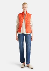 Gilet imbottito arancione brillante senza maniche su un maglione crema, abbinato a jeans in denim blu a gamba dritta e mocassini beige. Dettagli in tessuto a trama visibili.