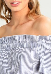 Top bez ramínek v modro-bílých vertikálních pruzích se zdobeným výstřihem a nařasenou texturou. Měkká, lehká látka.
