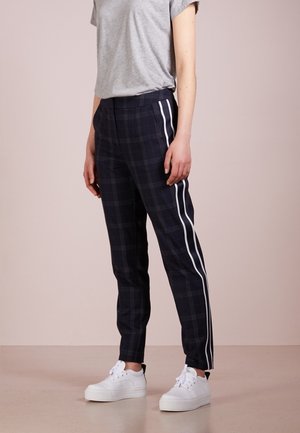 Persona que lleva pantalones oscuros de cuadros con rayas blancas laterales, camiseta gris de manga corta y zapatillas blancas, de pie sobre un suelo liso.