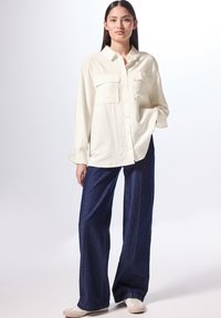 Camisa de manga larga color crema con dos bolsillos frontales, combinada con pantalones de mezclilla oscuros de pierna ancha y zapatillas de colores claros. Textura suave en toda la prenda.