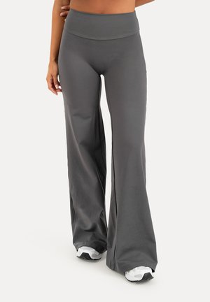 Donna che indossa pantaloni grigi a vita alta con gamba larga e sneakers bianche, in piedi con una mano appoggiata sul fianco.