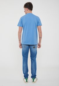 T-shirt di cotone blu a maniche corte, vestibilità rilassata, indossata con jeans azzurri e sneaker bianchi con accenti verdi.