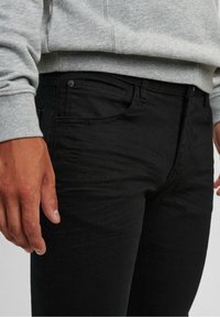 Vaqueros de mezclilla negra con un corte ajustado, que cuentan con un diseño clásico de cinco bolsillos y detalles de costura sutiles. Llevados con un sudadera gris.