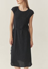Crna haljina do koljena s teksturiranim završetkom, kratkim rukavima i remenom u struku za prilagodljivu veličinu. Ima opušteni siluetu.
