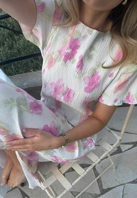 Γυναίκα καθισμένη ξυπόλητη σε λευκή μεταλλική καρέκλα, φοράει υφασμάτινο λευκό φόρεμα με ροζ λουλουδένια σχέδια, έξω σε πέτρινη πλατφόρμα.
