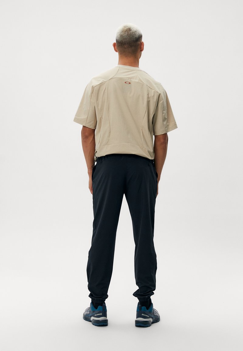 Beżowa koszula z krótkim rękawem o zaokrąglonym dolnym brzegu, zestawiona z granatowymi spodniami dresowymi. Model nosi niebieskie buty sportowe. Prosty, funkcjonalny design.