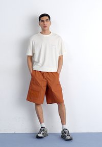 Camiseta de manga corta color beige claro, con un corte relajado, combinada con pantalones cortos cargo naranjas que tienen cuatro bolsillos. El modelo lleva zapatillas grises con calcetines blancos.