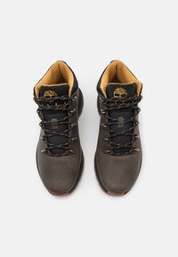 Botas de senderismo marrones que combinan materiales de cuero y textil, cordones redondos y una suela de goma con un diseño de banda; acentos amarillos en el interior.