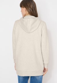 Femme aux longs cheveux bruns portant un sweat à capuche beige clair et un jean bleu, vue de dos sur fond blanc.