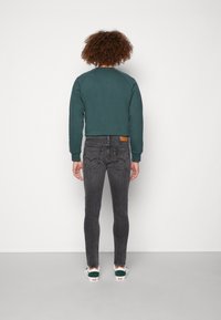 Donkergroene crewneck sweatshirt, grijze slim-fit jeans en witte sneakers met groene accenten. Zichtbaar logopatch op de tailleband van de jeans.