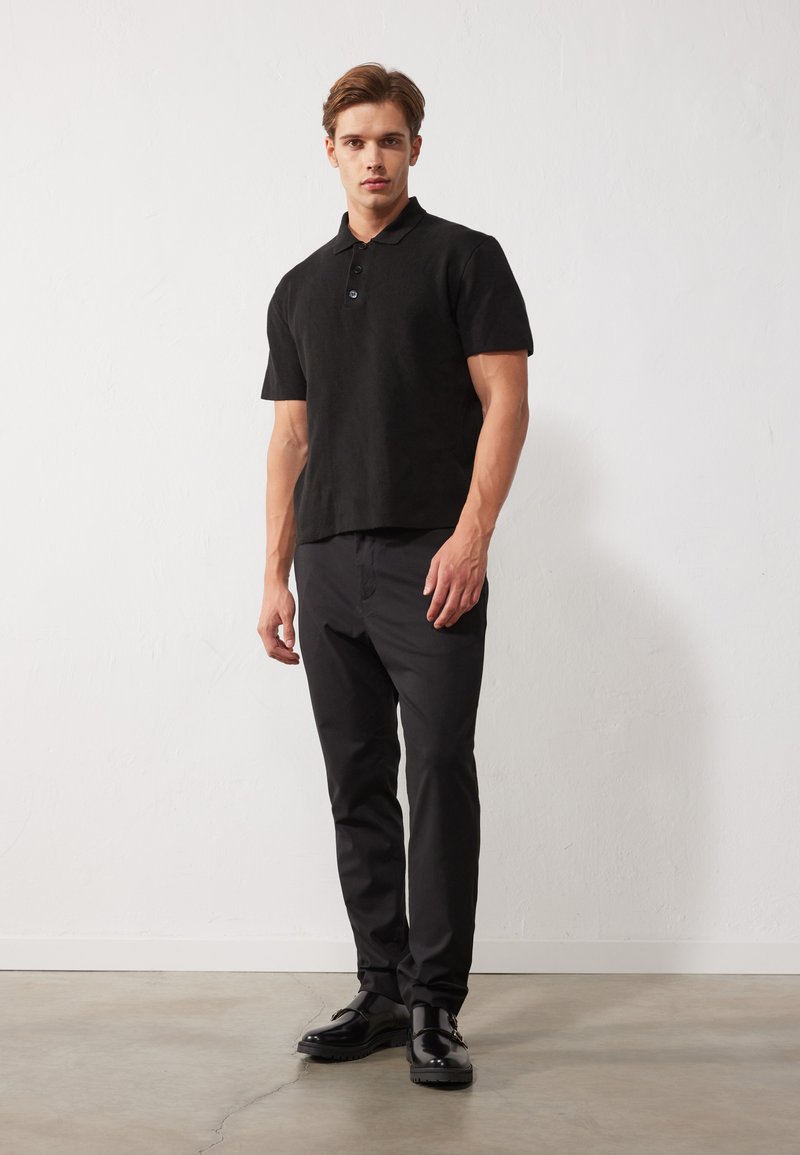 Fekete, rövid ujjú pólóing gallérral, három gombbal. Fekete, slim fit nadrággal és fényes fekete cipővel viselve.