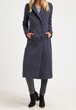 Vrouw draagt een lange marineblauwe dubbelgeknoopte jas met handen in de zakken, gecombineerd met grijze broek en zwarte enkellaarzen.
