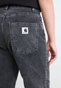 Sivé kordové nohavice s horizontálnou rib textúrou, s zadnou vreckom a bielym logom Carhartt. Minimalistický dizajn s vysokým pásom.