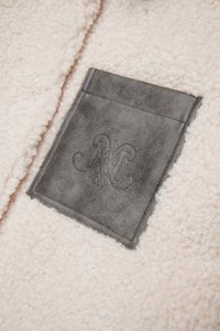 Lyserød fleece-stof med et firkantet mørkegråt læderpatch, der har en præget "M"-logo. Teksturen er blød og fluffy.
