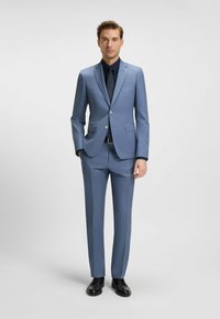 Jasnoniebieski garnitur z gładkiego materiału, z klapami w kształcie litery V, zapięciem na jeden guzik i prostymi spodniami. Czarna koszula i buty.