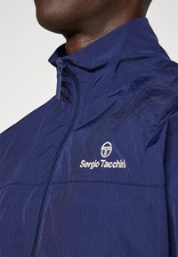 Marine lett jakke med høy krage, har frontglidelås og brodert "Sergio Tacchini" logo på venstre bryst.