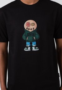 Černé tričko s potiskem karikatury s knoflíkovým okem a vyšitou pusou, oblečenou v zelené mikině, modrých kalhotách a teniskách.