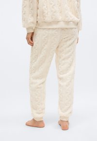 Άτομο που φοράει κρεμ παντελόνι από υφή φλις και αντίστοιχο πουλόβερ, όρθιο ξυπόλυτο μπροστά σε απλό ανοιχτό φόντο.