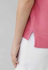 Lyserød strikket sweater med korte ærmer, der har sidesprækker og en ribbet kant. Båret over en hvid stof. Nærbillede af den nederste side og hånd.