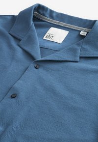 Polo bleu avec un tissu texturé, doté d'un col, de deux boutons noirs, et d'une étiquette portant la mention "NEXT EDIT". Col intérieur avec un design accentué.