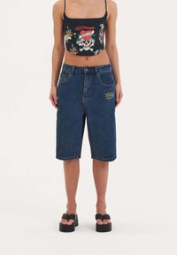 Black cropped top with graphic design, paired with blue denim Bermuda shorts featuring a yellow embroidered detail and side pockets. Black platform sandals.