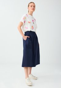 Jeune femme portant un t-shirt blanc à motif floral, une jupe midi bleu marine avec des poches et des baskets blanches, debout devant un fond clair uni.
