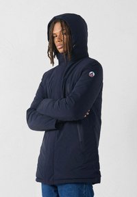 Giovane con dreadlocks che indossa un giubbotto invernale con cappuccio di colore blu navy, con le braccia incrociate, in piedi contro uno sfondo chiaro e semplice.