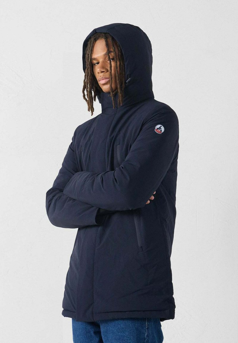 Giovane con dreadlocks che indossa un giubbotto invernale con cappuccio di colore blu navy, con le braccia incrociate, in piedi contro uno sfondo chiaro e semplice.