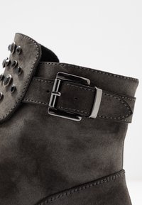 Bottine en daim gris avec une sangle à boucle, des clous noirs décoratifs et une texture lisse. Détails cousus le long des bords.