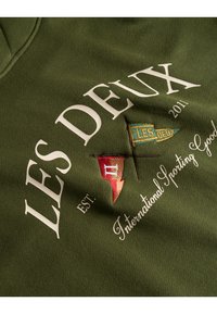 Felpa verde con dettagli ricamati. Presenta il testo "LES DEUX" e due bandiere colorate. Il materiale sembra morbido e testurizzato.