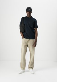 Camiseta navy con detalle de bolsillo, pantalones de pierna ancha beige, bolso negro de hombro y zapatillas blancas, llevada con una gorra negra. Diseño minimalista.