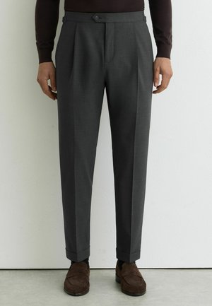 Mann trägt dunkelgraue, plissierte formelle Hose mit umgeschlagenen Säumen, braune Wildleder-Slipper und ein dunkles langärmeliges Hemd vor weißem Hintergrund.