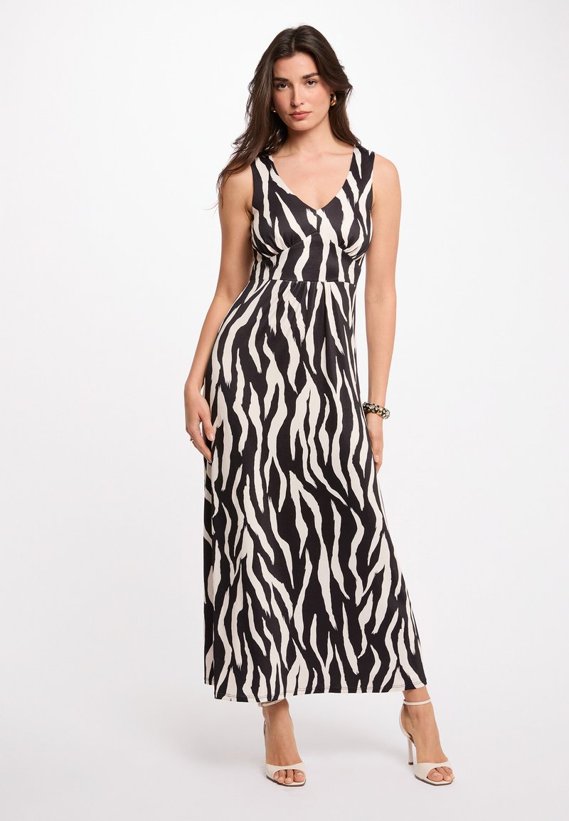 Mujer con un vestido maxi sin mangas de estampado de cebra en blanco y negro con escote en v y sandalias de tacón alto, posando contra un fondo blanco liso.