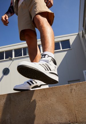 Osoba w beżowych szortach i białych butach Adidas stąpa po betonowej krawędzi przed nowoczesnym budynkiem pod czystym niebem.