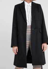 Kvinna klädd i en lång svart kappa över en vit skjorta med svarta vertikala ränder och högmidjade rutiga byxor i orangea och grå toner.