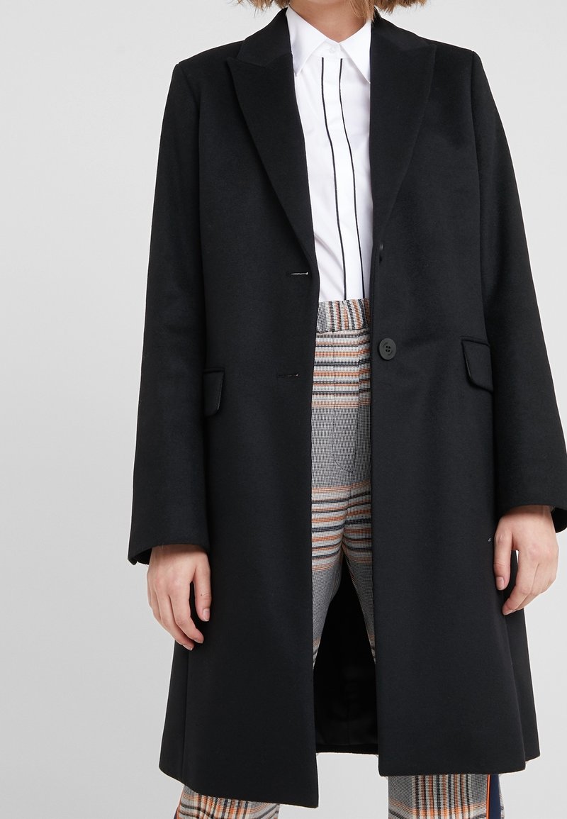 Kvinna klädd i en lång svart kappa över en vit skjorta med svarta vertikala ränder och högmidjade rutiga byxor i orangea och grå toner.