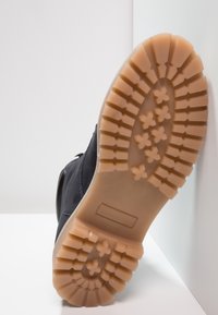 Bota negra con un upper de textil texturizado y una suela de goma que presenta un patrón de tracción robusto. La suela es de color marrón claro y tiene un perfil grueso.