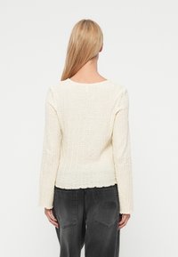 Haut à manches longues texturé de couleur crème avec un col rond et un ourlet légèrement festonné. Porté avec un pantalon gris foncé.