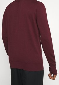 Pull en maille bordeaux avec des manches longues, un col rond et des poignets et ourlet côtelés. Texture lisse, design basique, sans motifs visibles.