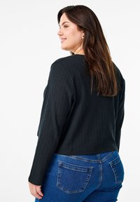 Črna rebrasta majica z dolgimi rokavi in krojeno zasnovo, v kombinaciji z modrimi teksas hlačami klasičnega pet-pocket sloga z zbledelim izgledom.