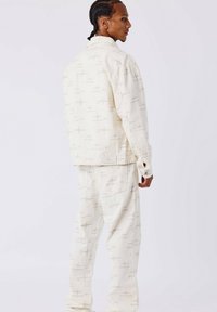 Ensemble de vêtements composé d'une veste et d'un pantalon en crème clair, fabriqué dans un tissu présentant un subtil motif beige de logos. Coupe décontractée avec des poches visibles et des détails en boutons.