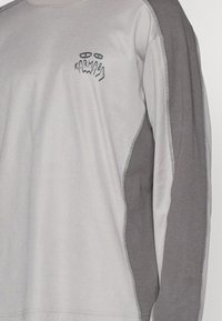 Long-sleeve shirt in light gray and dark gray panels, featuring a bold graphic print and visible stitching along the seams.