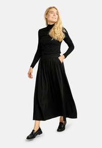 Czarna bluzka z długim rękawem ozdobiona błyszczącymi detalami, zestawiona z lekko opływającą czarną maxi spódnicą. Modelka nosi czarne loafersy, strój o luźnym kroju.