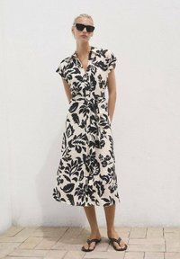 Robe à manches courtes et à boutons de couleur crème avec un motif floral noir, taille cintrée avec une ceinture nouée. Portée avec des sandales plates noires.