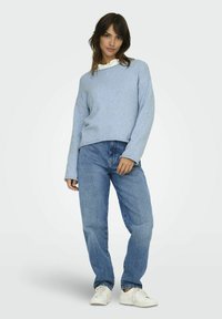 Sweater texturé bleu clair avec une coupe ample, porté par-dessus une chemise blanche à col, associé à un jean bleu droit et des baskets blanches.