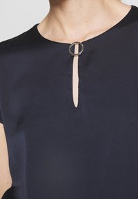 Marinblus med rund halsringning och korta ärmar, med en nyckelhålsöppning och en cirkulär metallaccent. Slät tygtextur.