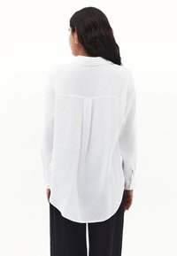Camicia bianca a maniche lunghe con tessuto testurizzato, yoke posteriore e orlo curvo. Abbinata a pantaloni neri larghi.