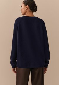 Navyblauer, übergroßer Pullover mit strukturiertem Strick, Raglanärmeln, Rundhalsausschnitt und lässiger Passform, kombiniert mit dunkelbraunen Hosen.